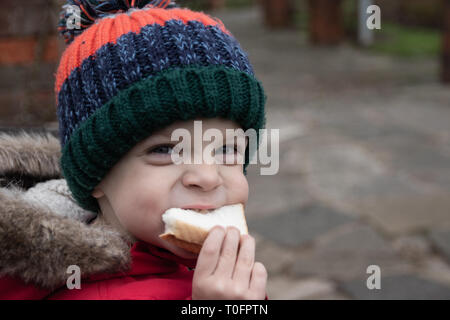 Piccolo Ragazzo di mangiare un panino indossando cappotto invernale e bobble hat Foto Stock