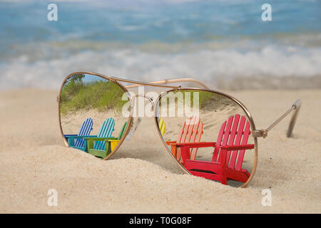 Coloratissima fila di sedie Adirondack riflessa in aviatore occhiali da sole in spiaggia di sabbia Foto Stock