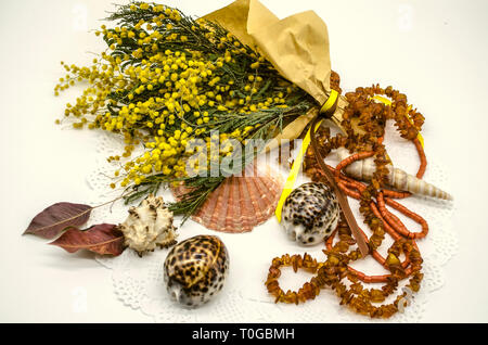 Un mazzo di fiori di giallo mimosa avvolto in carta ruvida accanto con conchiglie e perle di ambra e perle di corallo su un tracery tovagliolo bianco Foto Stock