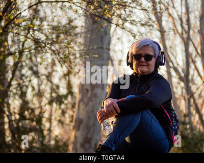 Un senior donna con i capelli bianchi per ascoltare musica con le cuffie e una bottiglia d'acqua in plastica in mano Foto Stock