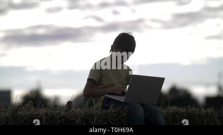 Ragazzo utilizza sul computer portatile sul pagliaio nel villaggio al tramonto Foto Stock