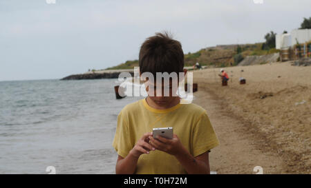 Ragazzo del viaggiatore a piedi sulla spiaggia e uso di smart phone in serata, chat, scrivere SMS. Utilizzando un palmare portatile dispositivo sulla vacanza estiva, internet. Foto Stock