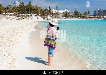 A piedi nudi donna in hat sulla spiaggia di scattare le foto del mare con lo smartphone. Vacanze estive, vacanze, viaggi e concetto di persone Foto Stock