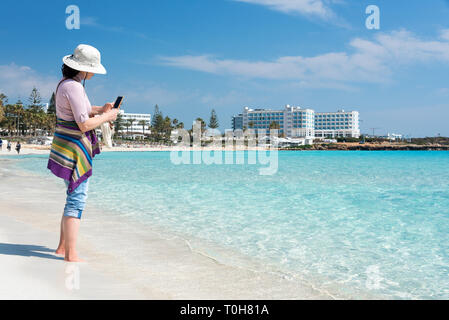 A piedi nudi donna in hat sulla spiaggia di scattare le foto del mare con lo smartphone. Vacanze estive, vacanze, viaggi e concetto di persone Foto Stock