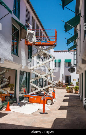 Due pittori che dipingono un stucco bianco edificio commerciale utilizzando un sollevatore a forbice e una scaletta mezzogiorno a Santa Barbara, California. Foto Stock