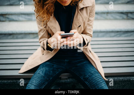 Close-up giovane donna che indossa il beige trench coat e jeans blu seduta sul banco grigio vicino a scalini in pietra utilizzando smart phone. Femmina mani dell'azienda mobile Foto Stock