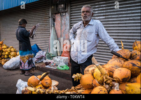 Fornitore di noce di cocco in piedi con la sua merce mentre un uomo legge una carta nelle vicinanze, Colombo, Sri Lanka Foto Stock