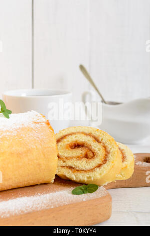 Delizioso rotolo di biscuit con marmellata di albicocche e una tazza di tè su un bianco tavolo di legno. Dessert per la prima colazione. Spazio di copia Foto Stock