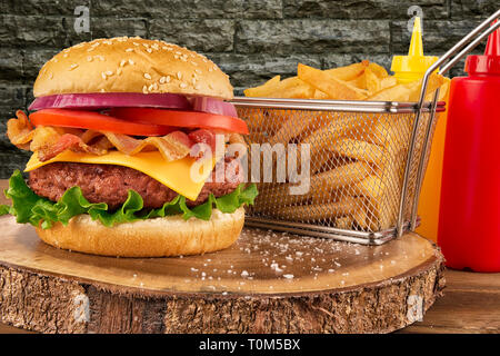 Cheeseburger con polpetta di carne di manzo e pancetta. Patate fritte nel cestello, ketchup e senape bottiglia in background. Isolato su un muro di mattoni. Real close up. Foto Stock