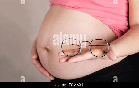 Gravidanza giovani tra i 30 e i 35 anni donna mantiene occhiali nella parte anteriore del suo ventre. Primo piano Foto Stock