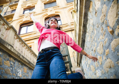 Felice giovane donna in rosa maglione ascoltando musica e saltare in strada. Basso angolo di visione Foto Stock