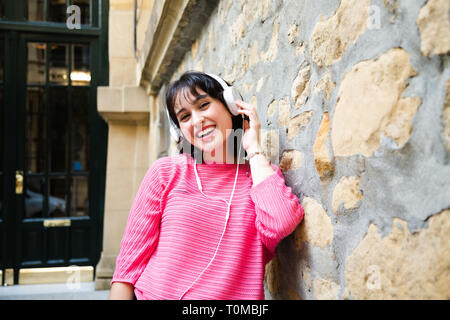 Felice bella ragazza in rosa maglione ascoltando musica in cuffie bianche appoggiata in una parete di strada. Foto Stock