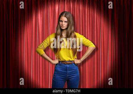 Giovani arrabbiati donna che indossa jeans e maglietta gialla sul palco rosso sfondo tende Foto Stock