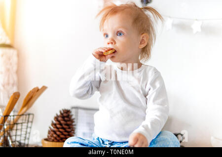 Poco carino toddler girl eating in casa cookie di avena e seduto sul tavolo della cucina mentre la sua sorella maggiore prepara la colazione. Famiglia di amore e di cura conc Foto Stock