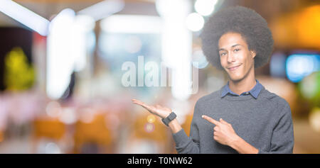 Giovani African American business man con capelli afro stupiti e sorridente alla fotocamera durante la presentazione con la mano e puntando con il dito. Foto Stock