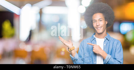 Giovane americano africano uomo con capelli afro stupiti e sorridente alla fotocamera durante la presentazione con la mano e puntando con il dito. Foto Stock