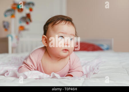 Ritratto di carino adorabile asiatica di razza mista bambina di quattro mesi di età che giace sulla pancia sul letto in camera da letto. Emozione naturale espressione faccia. Finestra giorno lig Foto Stock