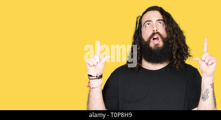 Giovane uomo con i capelli lunghi e la barba indossando metallo pesante abito nero meravigliato e sorpreso cercando e dispositivo di puntamento con le dita e le braccia sollevate. Foto Stock