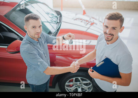 Acquirente e Rivenditore di stand e agitare ogni altre mani. Il venditore detiene le chiavi dall'auto. Entrambi di cercare su fotocamera. Sono vicino a auto rossa. Entrambi i giovani uomini a Foto Stock