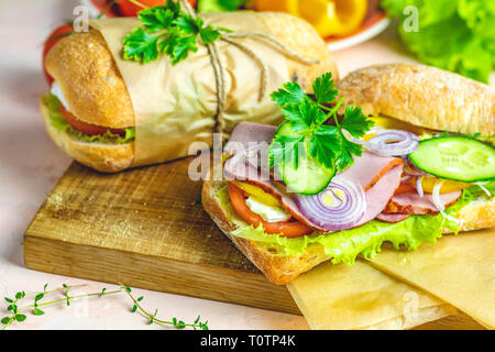 La ciabatta in sandwich con prosciutto e pomodoro, formaggio, pepe, cipolla e insalata sul tagliere di legno con ingredienti. Foto Stock