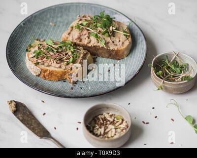 Due fette biscottate di anatra con prugne rillettes pate sul pane bianco con i germogli e diversi semi su una piastra di colore blu su sfondo bianco Foto Stock