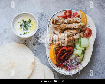 Pollo alla griglia su spiedini di metallo con delle verdure fresche sulla pita greca e la salsa in un recipiente su sfondo grigio. La cucina greca. Foto Stock