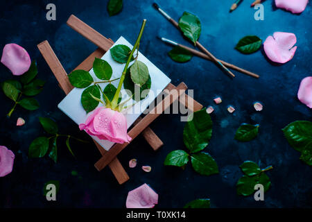 Rose fiore su un cavalletto pittore laici piana. Strumenti di pittura, pennelli e matite sul bagnato sfondo piovoso con copia spazio. La natura e il concetto di arte Foto Stock