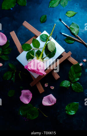 Rose fiore su un cavalletto pittore laici piana. Strumenti di pittura, pennelli e matite sul bagnato sfondo piovoso con copia spazio. La natura e il concetto di arte Foto Stock