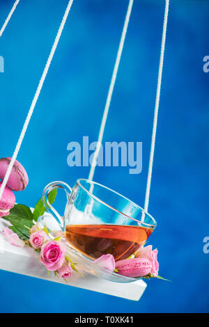 Rosa tea con macaron cookie e fiori di colore rosa su oscilla in movimento. Molla il concetto di energia su un vivace sfondo blu Foto Stock