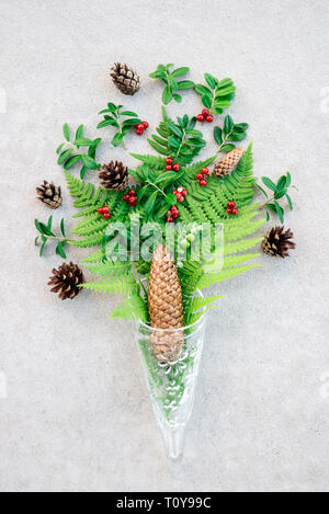 Cono in vetro con bosco selvatico piante, sul cemento grigio Sfondo. La felce, coni di conifere, cranberry foglie e frutti. Foto Stock