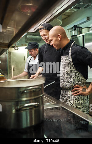 Ottimo lavoro. Allegro chef professionale guardando come i suoi due assistenti sono la cottura in un ristorante di cucina. La preparazione di alimenti Foto Stock