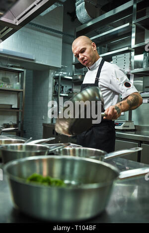 Molto caldo. Foto verticale di concentrato di chef in grembiule con parecchi tatuaggi sulle sue braccia tenendo una padella in un ristorante di cucina. Il processo di cottura Foto Stock