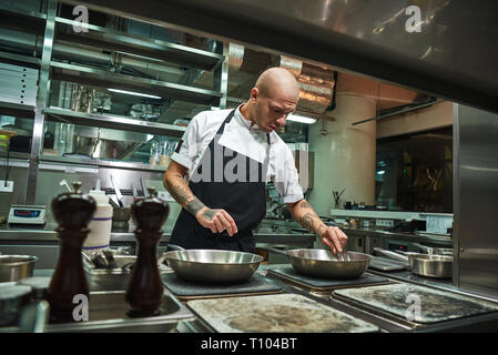 La preparazione di prodotti alimentari. Bello e fiducioso chef con parecchi tatuaggi sulle braccia per friggere ingredienti per il suo piatto in una cucina. Il processo di cottura Foto Stock