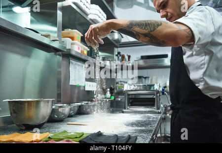 Slow motion. Bel giovane chef con il nero di tatuaggi sulle sue braccia versando la farina sul tavolo da cucina prima di fare la pasta. Cibo tradizionale Foto Stock