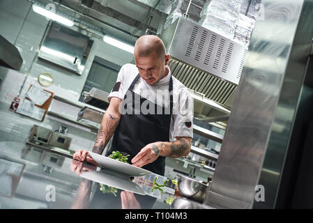 Concetto di cibo. Ritratto di bello chef professionale in nero grembiule decorare un insalata sulla piastra mentre si lavora in un ristorante di cucina. Pronto a servire Foto Stock