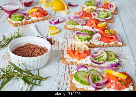 Pane croccante detto "Knäckebrot" aperto di fronte a sandwich con trota affumicata, morbida crema di formaggio, il cetriolo, la cipolla rossa, cosparsi di semi di lino in un bianco vecchio tavolo in legno, chiudi Foto Stock