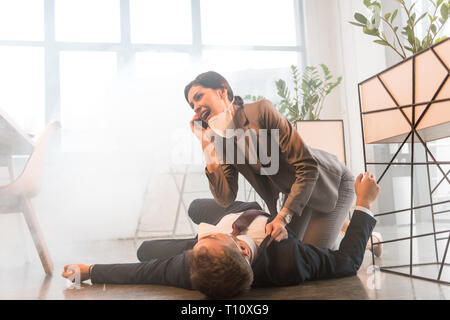 Terrorizzato imprenditrice in maschera parlando sullo smartphone vicino collega sdraiato sul pavimento in ufficio con il fumo Foto Stock