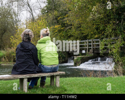 Vista posteriore di una madre e figlia in chat che indossa giacche mentre è seduto su una panchina lungo il fiume con un piccolo weir e cascata in background Foto Stock