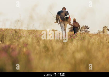 Vista posteriore del figlio e del padre andando per la pesca. Bassa angolazione di un uomo a piedi con un capretto a secco su un campo erboso. Foto Stock