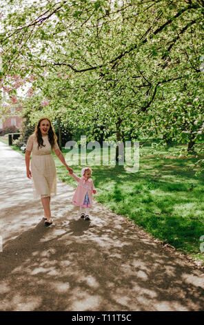 Bella donna in crema Abito in pizzo con la figlia in abito rosa passeggiate nel parco all'aperto albero verde, la molla. Famiglia sulla natura nel parco di Greenwich, Regno Unito Foto Stock