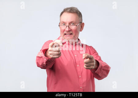 Emozionato senior Uomo in camicia rossa che indossa gli occhiali dita di puntamento in telecamera isolata su uno sfondo bianco. Egli è concordato con voi e felice con il vostro ch Foto Stock