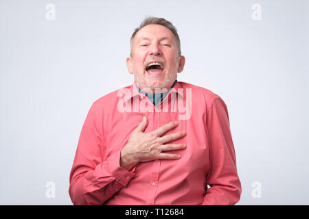 Senior Uomo in camicia rossa ride tenendo la mano sul petto. Positivo l'emozione del viso. Foto Stock