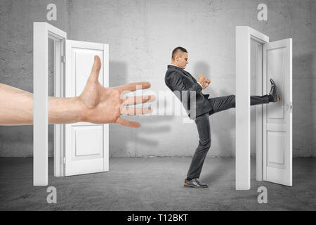 Imprenditore calci una porta ed una grossa mano che appaiono fuori da una porta aperta sul muro grigio sfondo Foto Stock