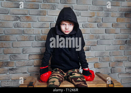 Little Boy boxer con capelli biondi medicazione in felpa nera indossando guanti da boxe in posa di un studio. Foto Stock