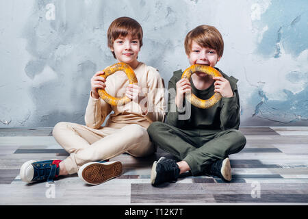 Due graziosi letti ragazzi seduti sul pavimento e felicemente mangiare bagel Foto Stock