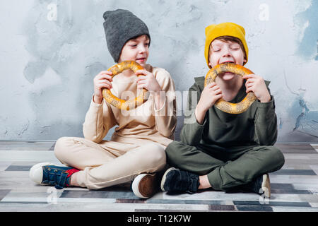 Due graziosi letti ragazzi seduti sul pavimento e felicemente mangiare bagel Foto Stock