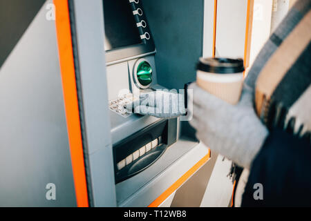 Close-up giovane donna premendo i pulsanti sulla tastiera ATM immettendo il PIN e di bere il caffè sulla strada di città nel giorno d'inverno. Foto Stock