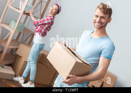 Giovane marito e moglie di passare a nuovo luogo donna in piedi vicino a ripiano guardando l uomo della scatola di contenimento cercando fotocamera a sorridere allegro Foto Stock