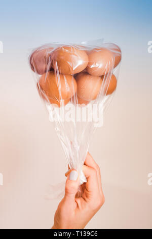 Le uova in un sacchetto di plastica capovolto come palloncini nella donna la mano sul multi colore blu e corallo chiaro sfondo rosa con luce morbida Foto Stock