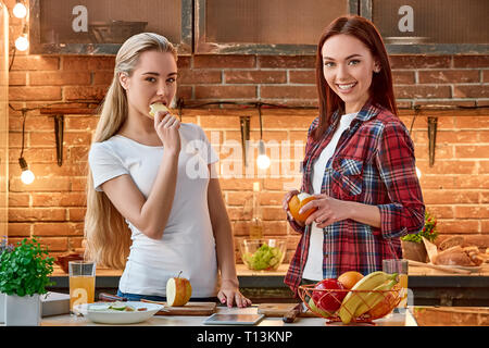 Ritratto di due donne attraenti, divertendosi, durante la preparazione di insalata di frutta. Essi sono pienamente coinvolti nel processo. Ragazza bionda in bianco T-shirt è guardando la telecamera mentre mangia apple slice. La sua scura con capelli amico in camicia a scacchi è in posa e la pelatura di un arancione. Vista frontale Foto Stock